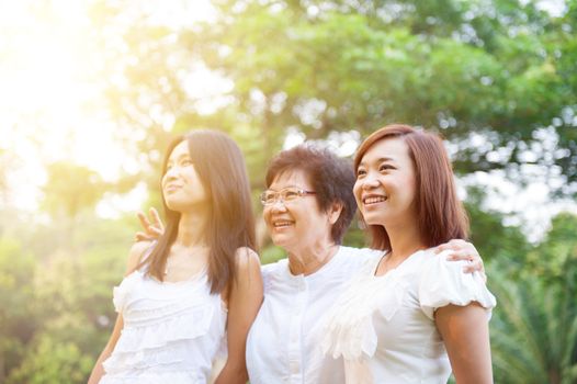 Portrait of Asian elderly mother and daughters looking away, senior adult woman and grown child. Outdoors family at nature park with beautiful sun flare.