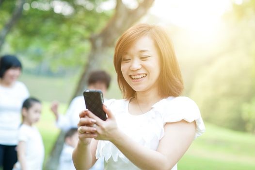 Asian woman texting using mobile phone at outdoor park, morning sun flare background.
