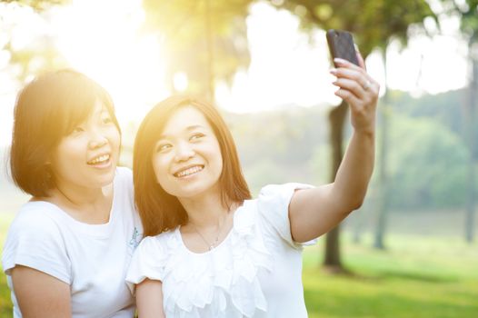 Young Asian females having fun at outdoor park, taking selfie using smartphone camera, sun flare background.