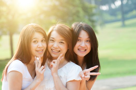 Group of young Asian females having fun at outdoor park, looking at camera showing peace hand sign, sisters or girlfriends, friendship concept, sun flare background.