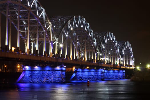 Railway bridge at night with white-blue illumination, reflection in the river