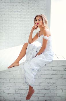 Beautiful girl in a white dress dreams of sitting on a brick wall