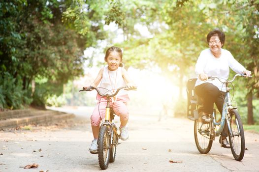 Portrait of active multi generations Asian family at nature park. Grandmother and granddaughter riding bicycle outdoor. Morning sun flare background.