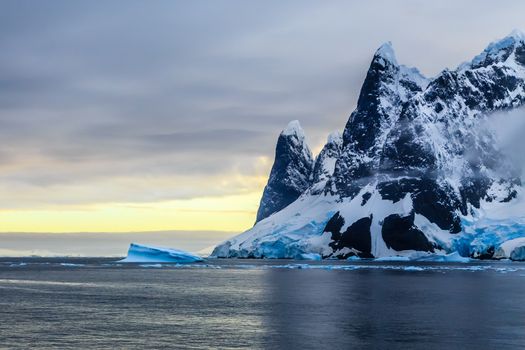 Sunset over cliffs, blue glacier and drifting iceberg with water surface in foreground, close to Argentine islands, Antarctica