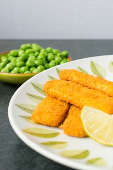 Breaded and Baked Fish Sticks, fingers  on a plate with a bowl of peas and a slice of lemon