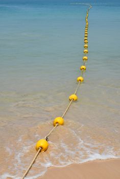 Chain of yellow polystyrene sea marker buoys with cable tow at sand beach and in blue sea water, perspective