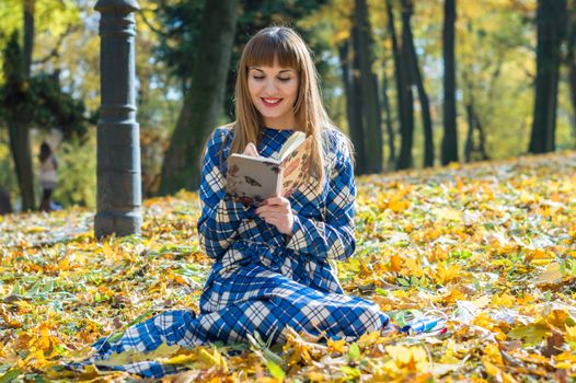 beautiful girl with long straight hair in a blue long dress reading a book in the autumn park