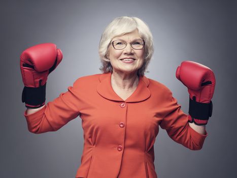 Smiling senior lady with raised hands in boxing gloves on gray background