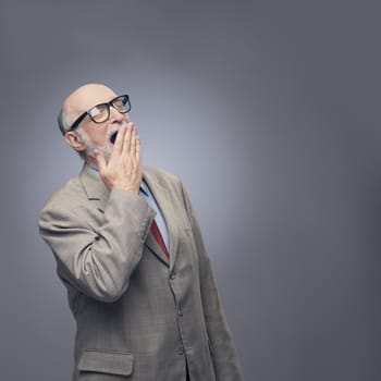 Yawning senior man in suit on gray background with copy space