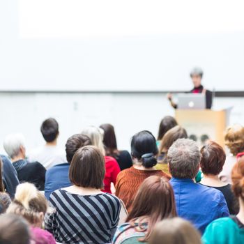 Business and entrepreneurship symposium. Female speaker giving a talk at business meeting. Audience in conference hall. Rear view of unrecognized participant in audience. Copy space on whitescreen.