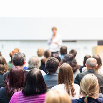 Business and entrepreneurship symposium. Female speaker giving a talk at business meeting. Audience in the conference hall. Rear view of unrecognized participant in audience. Square composition.