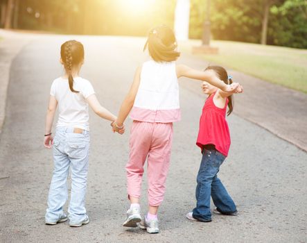 Rear view of Asian children holding hands walking at park. Little girls having fun outdoors. Morning sun flare background.