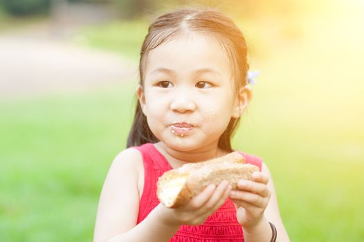 Portrait of Asian child eating sandwich at park. Little girl having fun outdoors. Morning sun flare background.