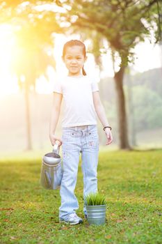 Portrait of cute Asian kid watering plant outdoors. Little girl having fun at nature park. Morning sun flare background.