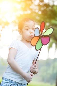 Portrait of cute Asian child playing windmill toy outdoors. Little girl having fun at nature park. Morning sun flare background.
