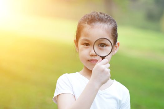Portrait of Asian child with magnifier glass exploring nature at park. Little girl having fun outdoors. Morning sun flare background.
