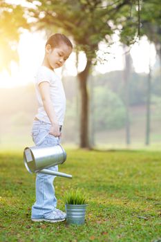 Portrait of active Asian child watering plant outdoors. Little girl having fun at nature park. Morning sun flare background.