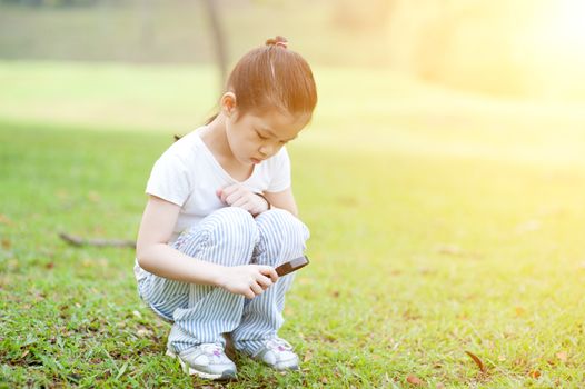Portrait of Asian school kid with magnifier glass exploring nature at park. Little girl having fun outdoors. Morning sun flare background.