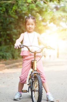 Portrait of active Asian child riding bike outdoors. Little girl having fun at nature park. Morning sun flare background.