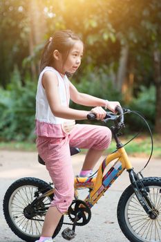 Portrait of active Asian child cycling outdoors. Little girl having fun at nature park. Morning sun flare background.