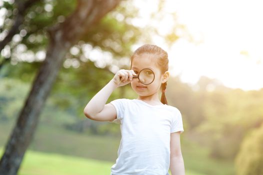 Portrait of Asian girl with magnifier glass exploring nature at park. Little child having fun outdoors. Morning sun flare background.