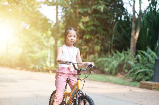 Portrait of active Asian child riding bicycle outdoors. Kid having fun at nature park. Morning sun flare background.