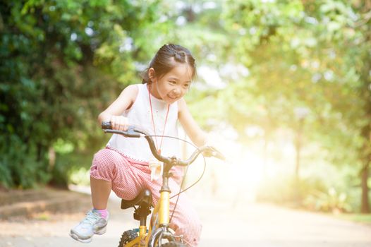 Portrait of active Asian kid biking outdoors. Little girl having fun at nature park. Morning sun flare background.