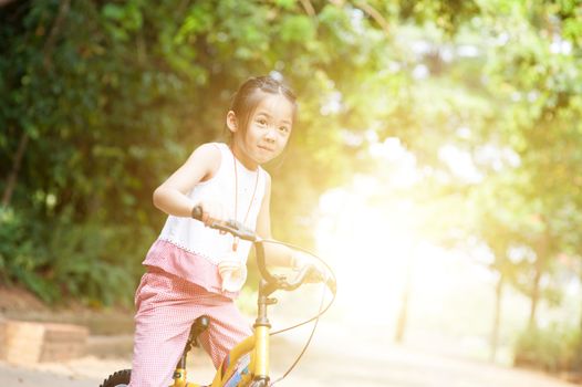 Portrait of active Asian child riding bicycle outdoors. Little girl having fun at nature park. Morning sun flare background.