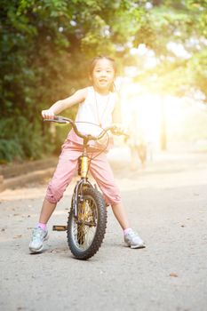 Portrait of active Asian child biking outdoors. Little girl having fun at nature park. Morning sun flare background.