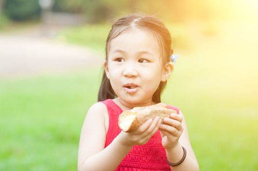 Portrait of Asian kid eating sandwich at park. Little girl having fun outdoors. Morning sun flare background.