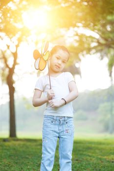 Portrait of cute Asian kid playing windmill toy outdoors. Little girl having fun at nature park. Morning sun flare background.