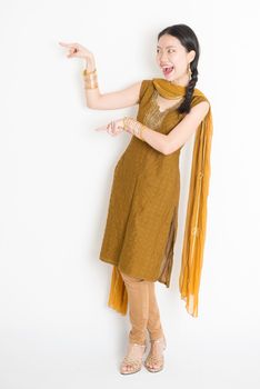 Portrait of young mixed race Indian Chinese girl in traditional punjabi dress fingers pointing at copy space, full length standing on plain white background.