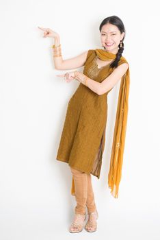 Portrait of young mixed race Indian Chinese woman in traditional punjabi dress fingers pointing at copy space, full length standing on plain white background.