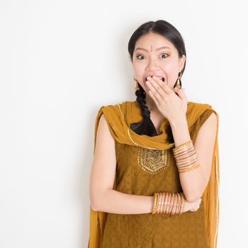 Portrait of shocked mixed race Indian Chinese girl in traditional punjabi dress covering her mouth, surprised emotion, standing on plain white background.