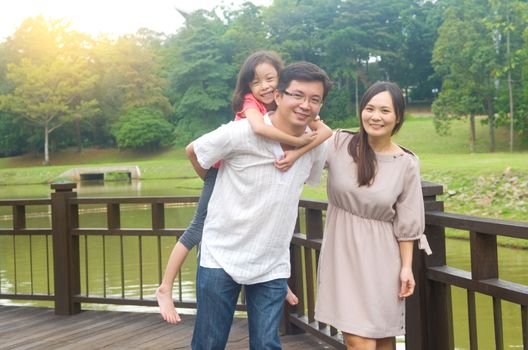 Happy Asian family outdoor. Father piggybacking his daughter walking in garden park with wife. Healthy lifestyle.