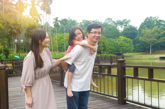 Happy Asian family outdoor. Father piggybacking his daughter walking in garden park with wife. Healthy lifestyle.