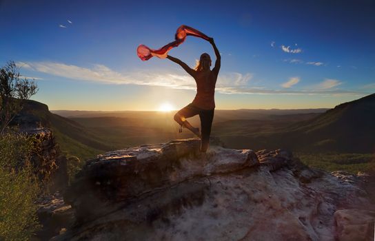 A woman balancing on one leg in mountain landscape, she has her arms outstretched holding sheer flowing fabric backlit by the setting sun moving with the breeze