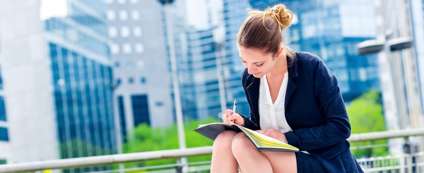 dynamic young executive girl taking notes on her agenda, outside. Symbolizing a job search or a trade of outsourcing