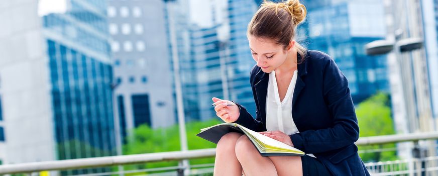 panoramic view on dynamic young executive girl taking notes on her agenda, outside. Symbolizing a job search or a trade of outsourcing