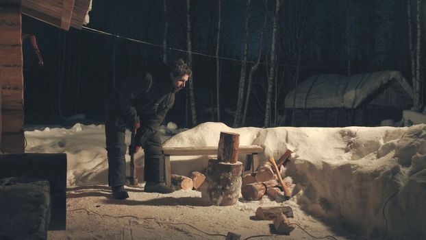 Young man chopping wood in the yard. Winter evening. Wide shot. 4K