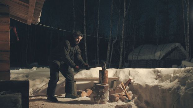 Young man chopping wood in the yard. Winter evening. Wide shot. 4K