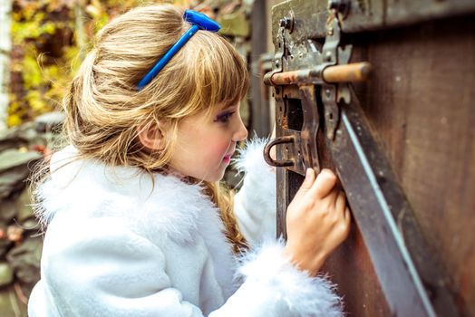 Side view of an little beautiful girl with long wavy hair in a blue silk dress in the scenery of Alice in Wonderland looking into the keyhole of the gate.