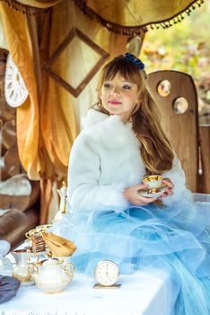 Front view of an little beautiful girl in the scenery of Alice in Wonderland holding a cup of tea at the table in the autumn park.