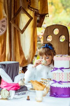 Front view of an little beautiful girl in the scenery of Alice in Wonderland drinking a tea at the table in the drinking tea in the autumn park.