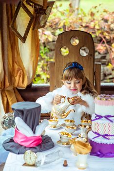 Front view of an little beautiful girl in the scenery of Alice in Wonderland pouring tea into a cup at the table in the autumn park.