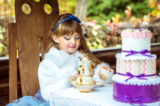 Front view of an little beautiful girl in the scenery of Alice in Wonderland holding a cup of tea at the table in the autumn park.