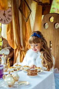 Front view of an little beautiful girl in the scenery of Alice in Wonderland holding a cup of tea at the table in the autumn park.