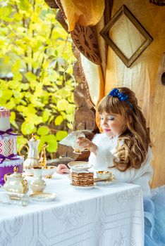 Front view of an little beautiful girl in the scenery of Alice in Wonderland holding a cup of tea at the table in the autumn park.