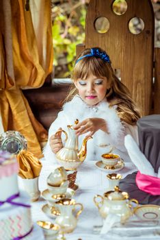 Front view of an little beautiful girl in the scenery of Alice in Wonderland pouring tea into a cup at the table in the autumn park.