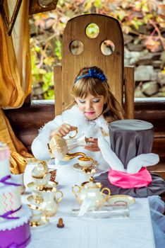 Front view of an little beautiful girl in the scenery of Alice in Wonderland pouring tea into a cup at the table in the autumn park.